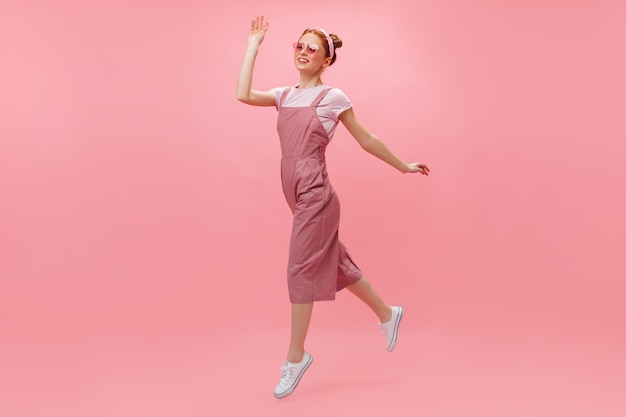 Free photo red-haired woman in overalls and glasses joyfully moves on pink background.