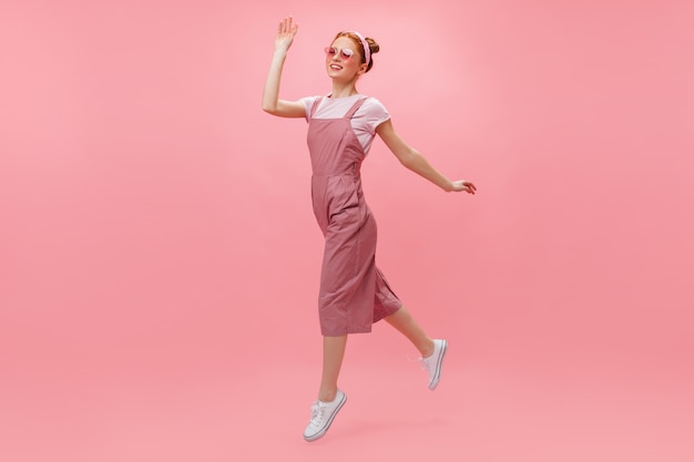 Red-haired woman in overalls and glasses joyfully moves on pink background.