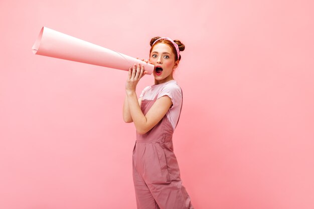 Red-haired woman in pink jumpsuit shouts loudly into horn on isolated background.