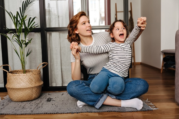 Red-haired woman in striped T-shirt hugs her daughter and plays with her sitting on floor in living room.