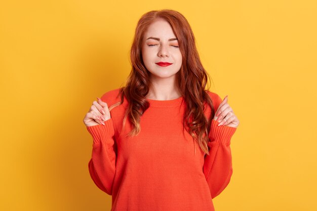 Red haired woman with closed eyes clenched fists, making wish, wearing orange sweater, standing isolated