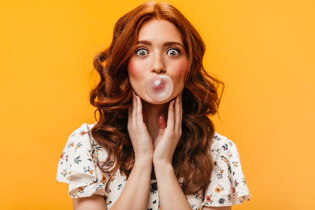 Free photo red-haired woman with green eyes in astonishment looks at camera on orange background. woman in white t-shirt makes bubble of gum.