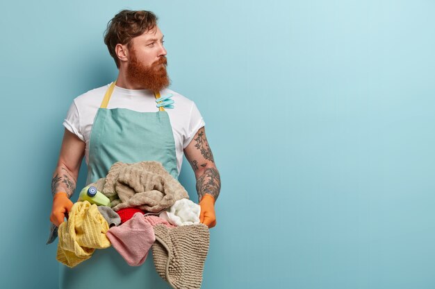 Red haired young man overstained with housekeeping, holds basin with pile of laundry, wears casual t shirt and apron, looks away, isolated over blue wall, has thoughtful look, focused away