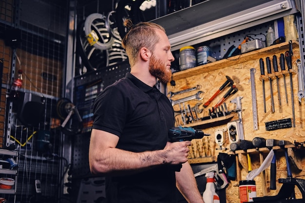 Рыжий бородатый велосипедный механик держит отвертку над подставкой для велосипедных инструментов в мастерской.