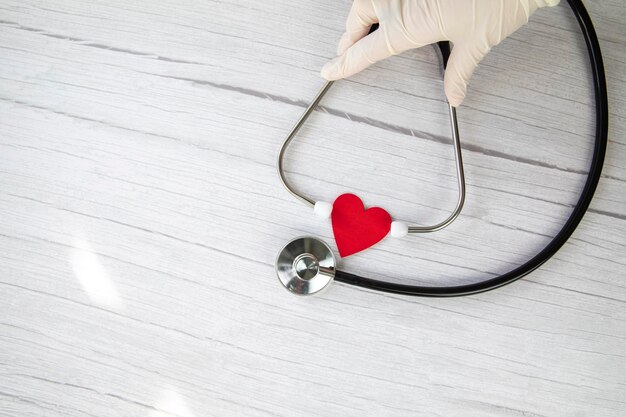 Red heart and white protective medical gloves holds a stethoscope on a white wooden background conce...