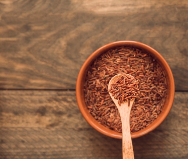 Red jasmine rice grains on spoon over the bowl against wooden backdrop