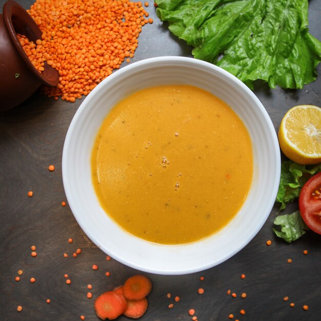 Суп из красной чечевицы в миске с морковью, листьями салата, лимоном, помидорами, сырой чечевицей