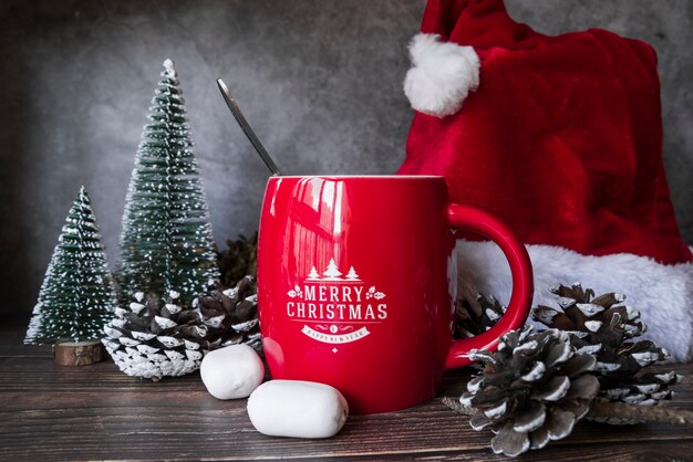 Red mug near Christmas hat and little fir trees 