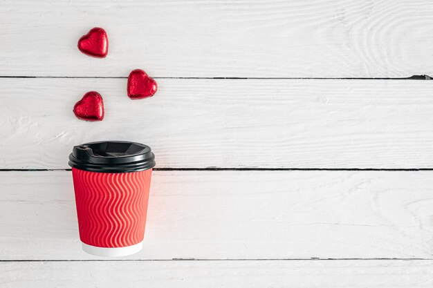 Red paper cup and heartshaped candies on a white wooden background