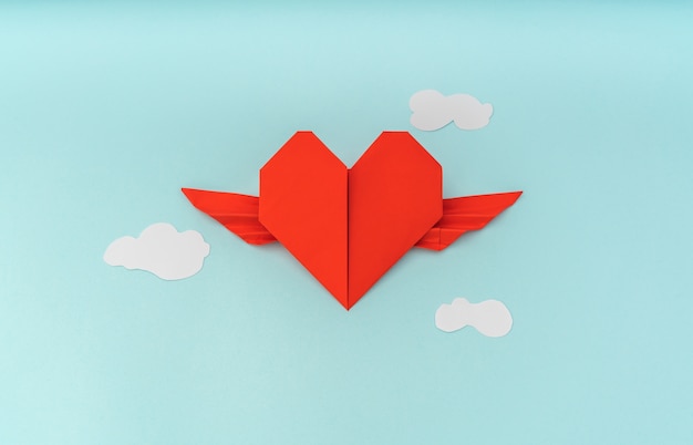 Red paper origami heart with wings and cloud on blue background
