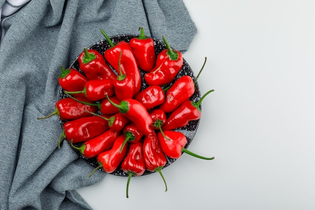 Red peppers in a plate on white and textile wall, top view.