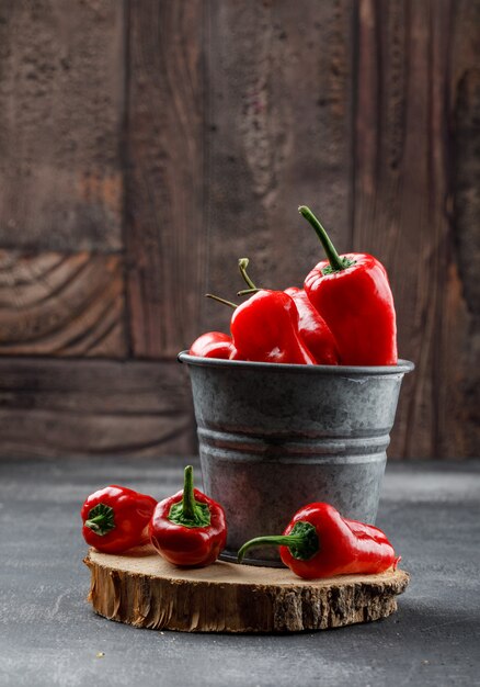 Red peppers with wooden piece in a mini bucket on grungy and stone tile wall, side view.