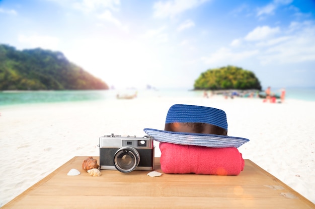 Red pink tower, blue hat, old vintage camera and shells over wooden floor on sunshine blue sky and ocean background