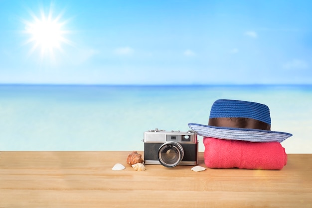 Red pink tower, blue hat, old vintage camera and shells over wooden table on sunshine blue sky and ocean background