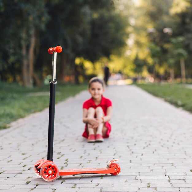Красный толчок скутер перед девушкой, сидящей на дорожке в парке