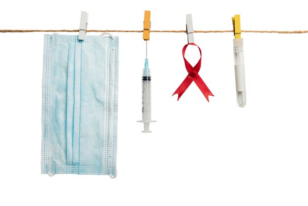 白い背景のロープにぶら下がっている赤いリボン、注射器、フェイスマスク。 HIVエイズリボンの認識