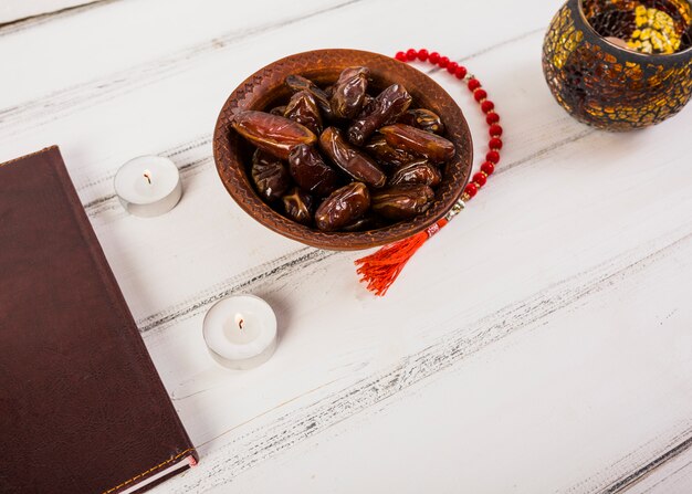 Red rosary beads with bowl of juicy dates and candles on white wooden table