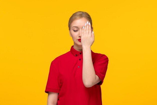 黄色の背景に赤いシャツで片目を覆う赤いシャツの日のブロンドの女性