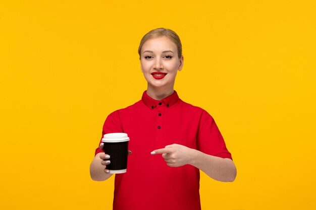 빨간 셔츠 날 노란색 배경에 빨간 셔츠에 커피 컵을 가리키는 귀여운 소녀