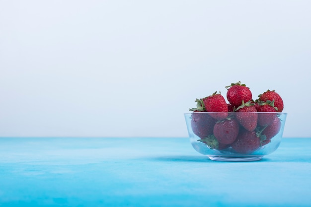 Red strawberries in a glass cup on blue, angle view.