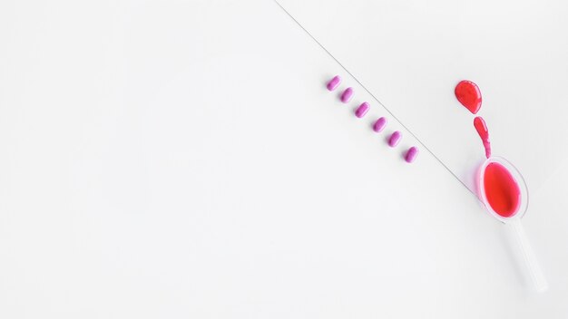 Red syrup in spoon with row of pills on white background