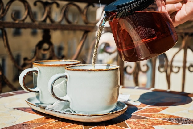 ガラスのティーポットに入った赤茶をカップに注ぐ古いベランダでの朝のお茶の時間夏休みのお茶会の雰囲気休息とリラクゼーションカップに選択的な焦点を当てる