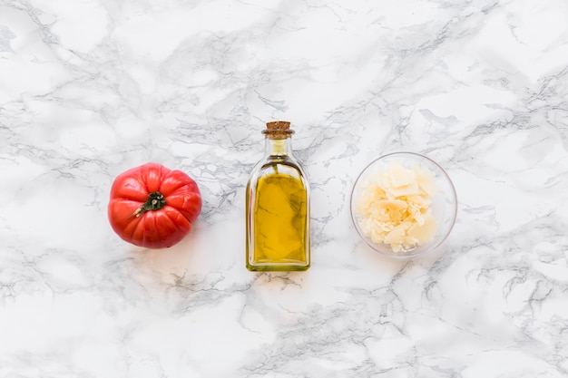 Red tomato with olive oil bottle and grated cheese in bowl on white marble backdrop