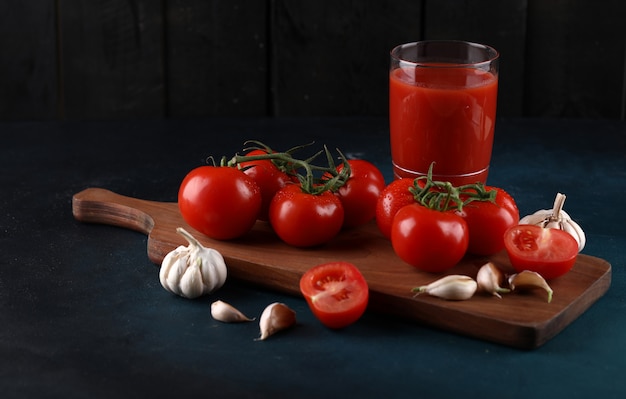 Red tomatoes and garlic gloves on the wooden board with a glass of juice on the blue background.