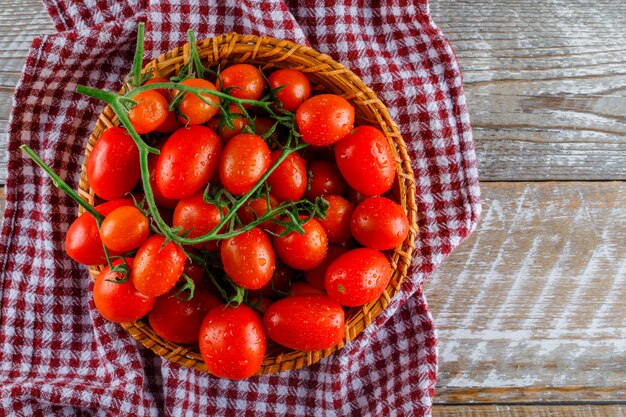 Red tomatoes in a wicker basket on wooden and kitchen towel, flat lay.