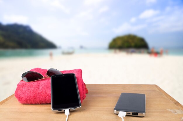 Red tower, sun glasses, mobile charging with power bank over wooden table on blur beach sand and blue sky background.
