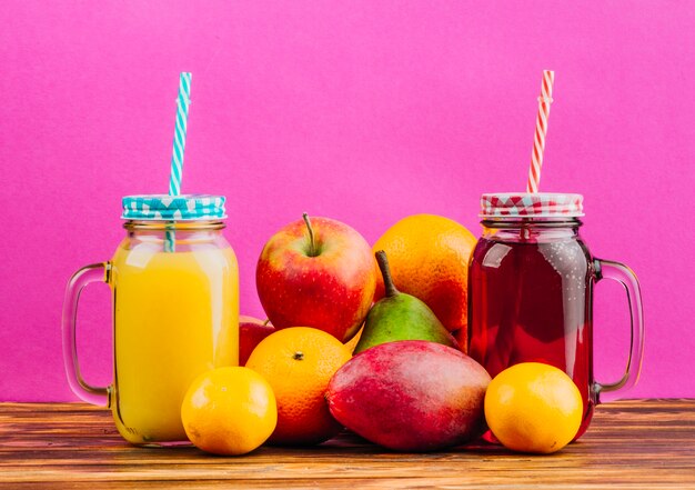 Red and yellow juice mason jars with drinking straws and fresh fruits against pink background