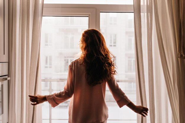 Redhaired girl touching curtains Indoor photo of caucasian young lady standing near window