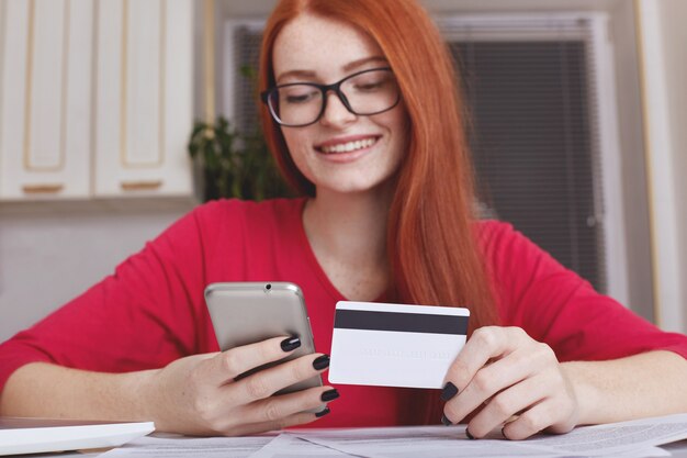 Рыжеволосая довольно женская модель в очках держит смартфон и кредитную карту