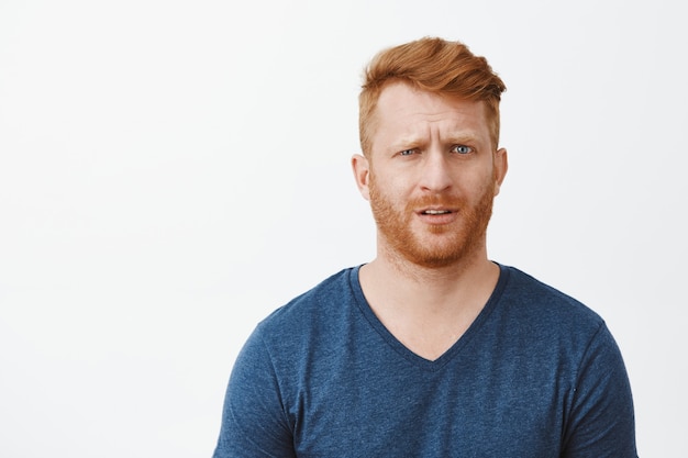 Redhead adult man in stupor cannot understand what happening, feeling clueless and questioned, lifting eyebrow and staring confused, standing uncertain over gray wall