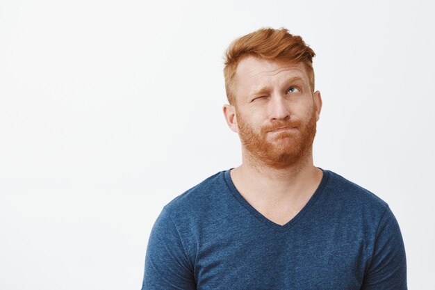 Redhead attractive male with bristle in casual blue t-shirt, pursing lips making hmm sound, closing one eye and looking up, thinking or making decision