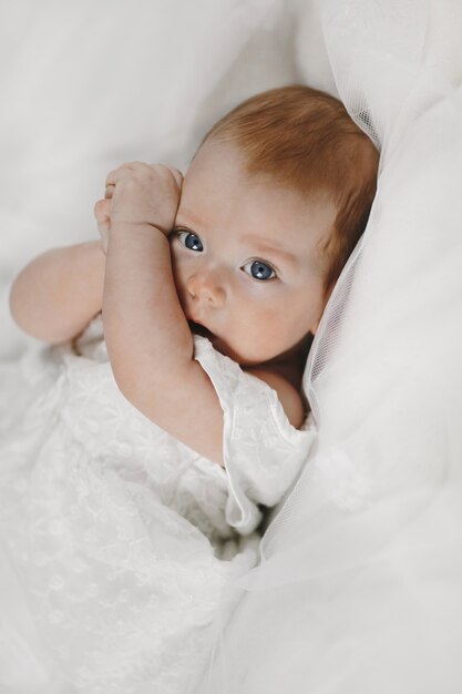 Redhead baby girl is lying on the white blanket with big clear blue eyes, and covering face with tiny arms