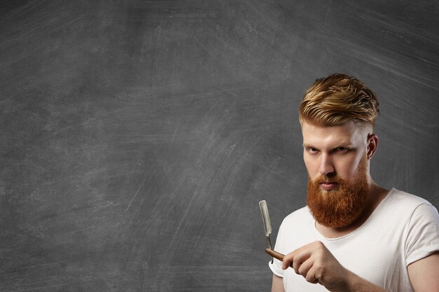 Redhead barber with stylish haircut and hipster beard holding his barbershop accessory - old-fashioned straight razor.