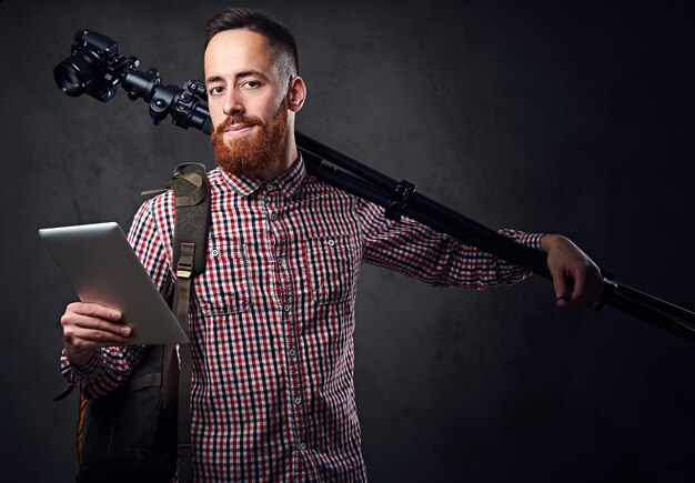 Redhead bearded hitchhiker holds tablet PC and a camera tripod.