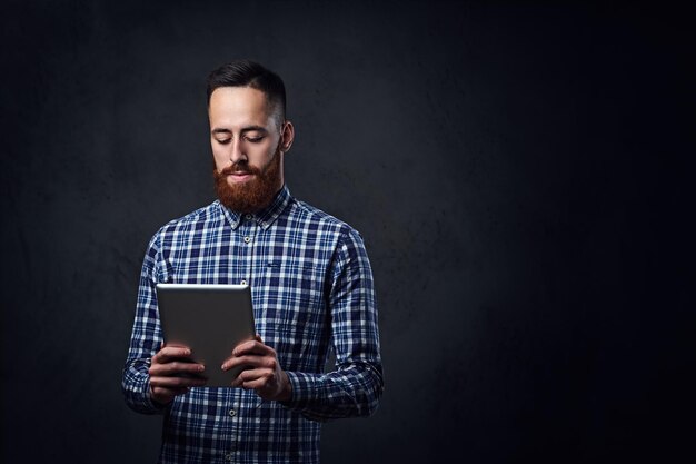 Redhead bearded male dressed in a blue fleece shirt holds tablet PC on grey background.