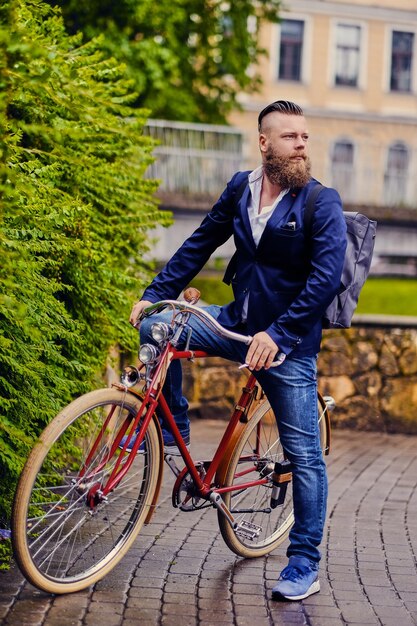 빨간 머리 수염 난 남성은 공원에서 복고풍 자전거에 파란색 재킷과 청바지를 입고.