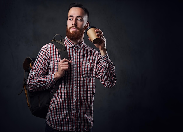 フリースシャツを着た赤毛のひげを生やした男性は、バックパックを保持し、コーヒーカップをテイクアウトします。