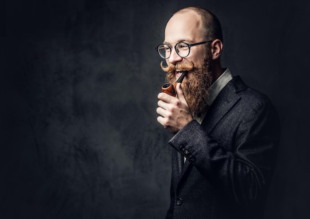 Redhead bearded male dressed in a suit and eyeglasses smoking tradition pipe over dark grey background.