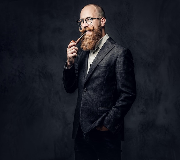 Redhead bearded male dressed in a suit and eyeglasses smoking tradition pipe over dark grey background.