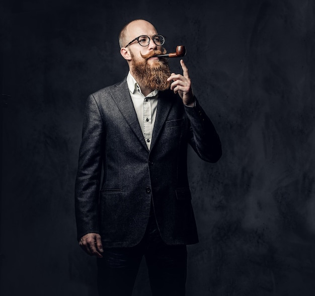 Redhead bearded male dressed in a suit and eyeglasses smoking tradition pipe over dark grey background.