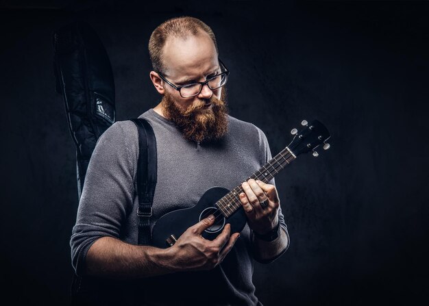 Redhead bearded male musician wearing glasses dressed in a gray t-shirt playing on a ukulele. Isolated on dark textured background.