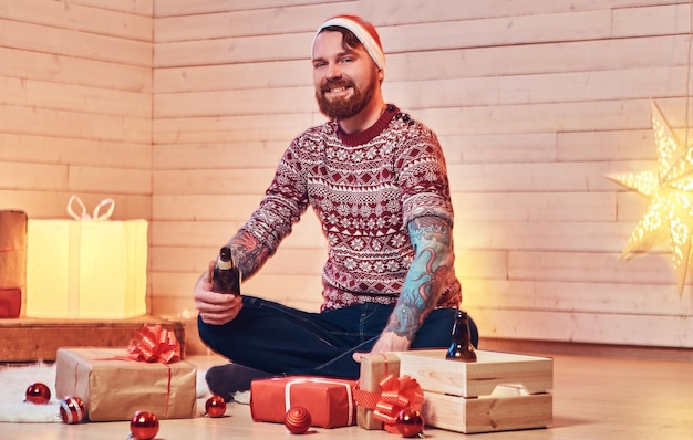 Redhead bearded male in Santa hat, drinks craft beer in a room with Christmas decoration.