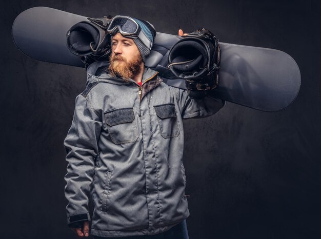 Redhead bearded snowboarder in a winter hat and protective glasses dressed in a snowboarding coat posing with snowboard at a studio, looking away. Isolated on a gray background.