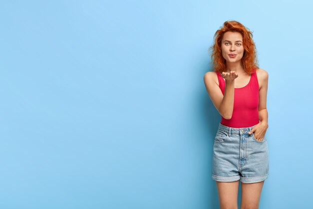 Redhead beautiful young woman sends air kiss, looks passionately, expresses love to followers, dressed in red vest and denim shorts, models against blue wall. Mwah and all my love to you.