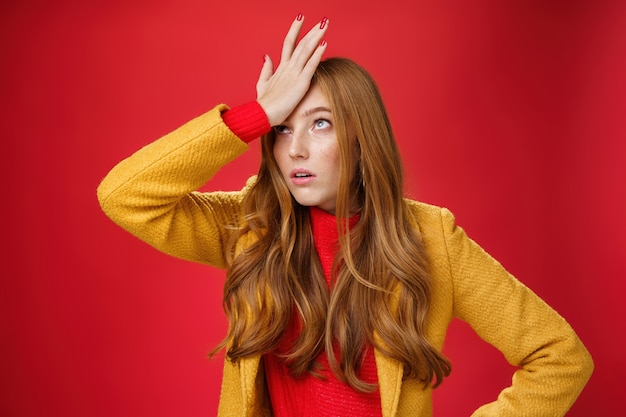 Free photo redhead businesswoman making facepalm gesture with hand on forehead rolling eyes up from annoyance and irritation as being shocked with how dumb client is sighing bothered over red wall, tired.