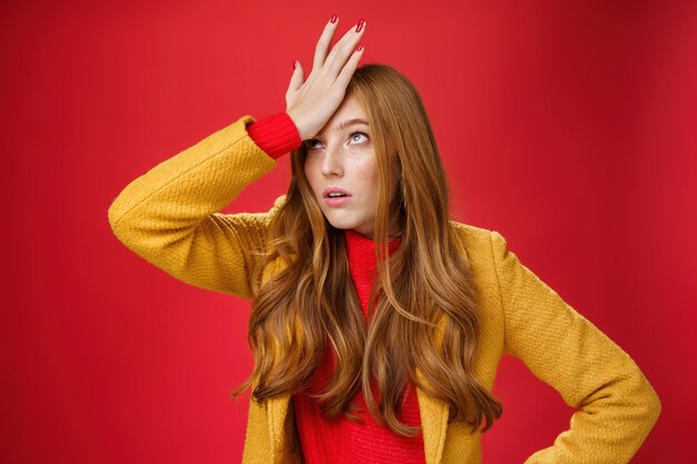 Redhead businesswoman making facepalm gesture with hand on forehead rolling eyes up from annoyance and irritation as being shocked with how dumb client is sighing bothered over red wall, tired.